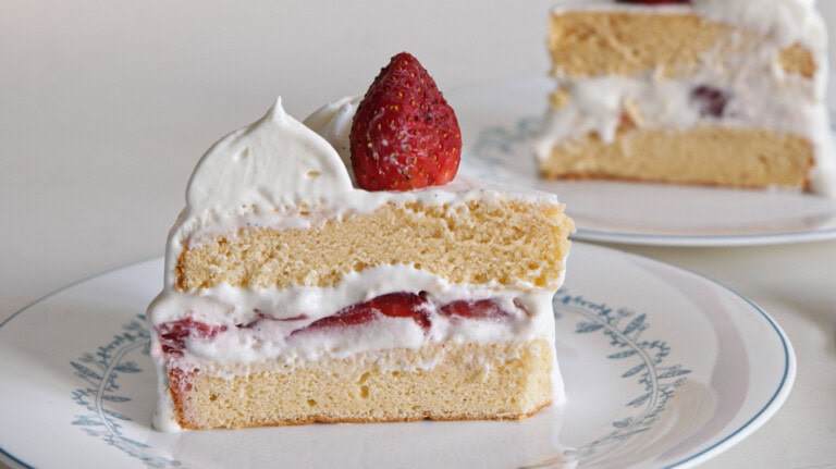 Two slices of sponge cake layered with whipped cream and fresh strawberries are served on white plates. Each slice is topped with a dollop of whipped cream and a whole strawberry. A gold fork is placed beside one of the plates.