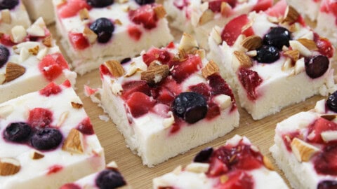 A close-up of several square pieces of frozen yogurt bark lying on a wooden surface. Each piece is topped with a mixture of blueberries, strawberries, and chopped nuts. The bark has a creamy white base.