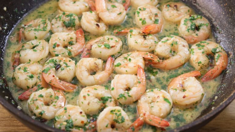 A pan filled with cooked shrimp in a garlic butter sauce, garnished with chopped parsley. The shrimp are evenly distributed and have a light pink color, indicating they are fully cooked. The sauce appears creamy and is sprinkled with herbs and red pepper flakes.