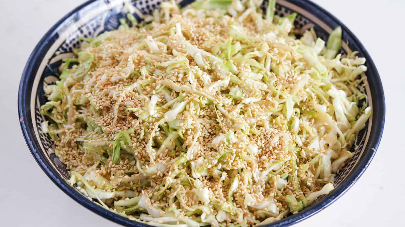 A bowl filled with a cabbage salad topped with sesame seeds. The shredded cabbage is mixed with a dressing, and the salad appears fresh and vibrant. The bowl is blue with a decorative pattern on the rim.