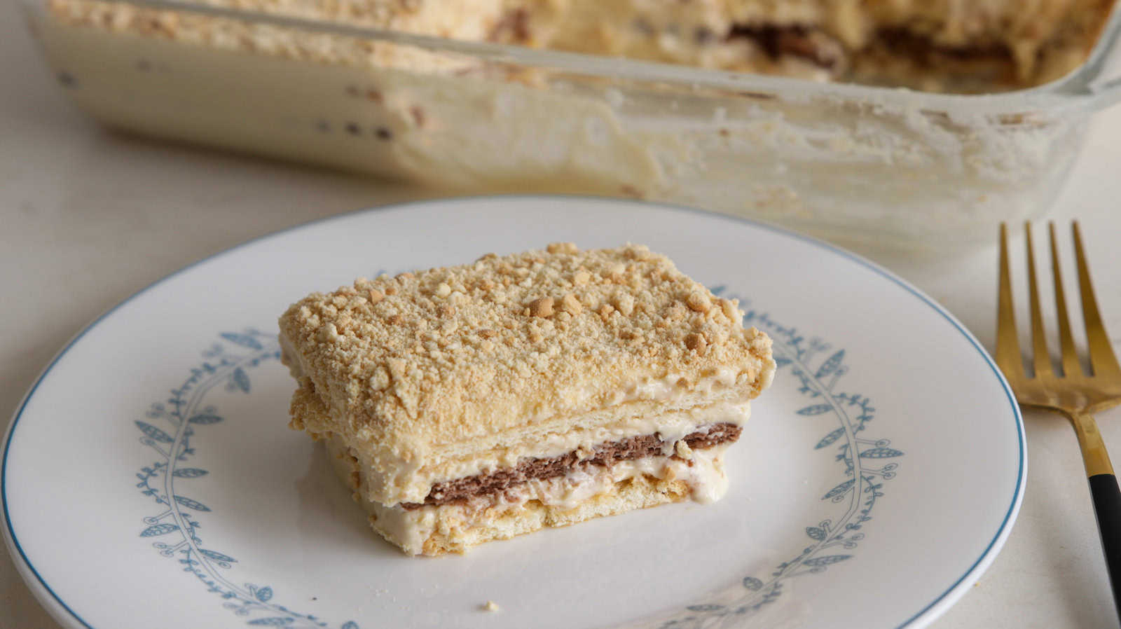 A slice of layered dessert consisting of cream and chocolate layers sits on a white plate with a blue floral border. The dessert has a crumbly top layer. In the background, there is a glass dish containing the remaining dessert. A fork is placed beside the plate.