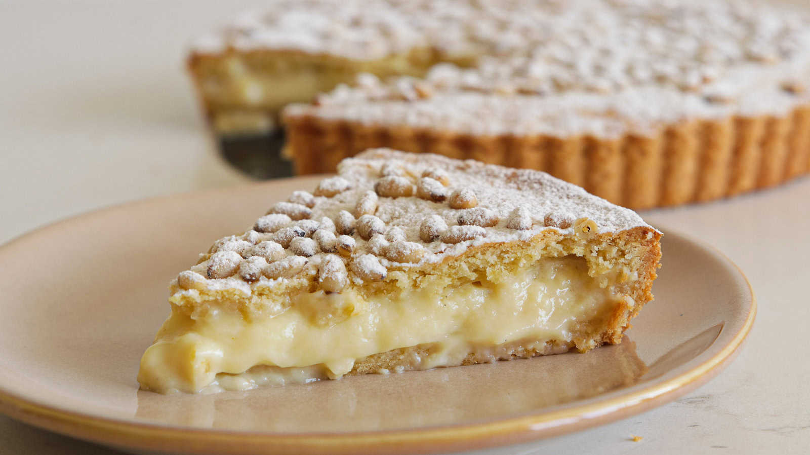 A close-up of a slice of torta della nonna on a beige plate. The pie has a golden brown crust and is topped with powdered sugar. In the background, the whole pie, with a conspicuously missing slice, is partially visible.