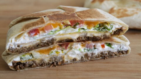 Homemade Crunchwrap Supreme Recipe A close-up of a halved grilled crunhwrap suprme with ground beef, lettuce, tomato, cheese, and sour cream, stacked on a wooden surface.