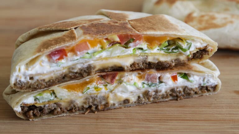 A close-up of a halved grilled crunhwrap suprme with ground beef, lettuce, tomato, cheese, and sour cream, stacked on a wooden surface.
