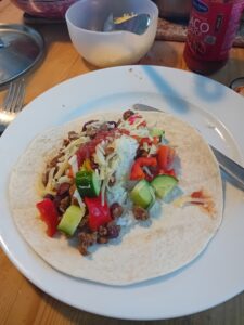 A soft tortilla wrap is filled with cooked minced meat, white rice, shredded cheese, chopped red bell peppers, cucumber pieces, and a drizzle of sauce. The wrap sits on a white plate with a fork and knife beside it. In the background, there's a bottle of taco sauce and a bowl with cheese.