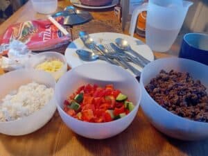 A wooden table with bowls of cooked rice, ground beef, and diced red bell peppers with cucumbers. A small bowl with shredded cheese, spoons on a white plate, a plastic jug with water, and a pack of tortillas are also on the table.