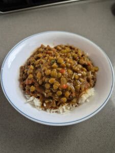 A bowl of white rice topped with a lentil curry containing green lentils, tomatoes, and herbs, served in a white bowl on a gray countertop.