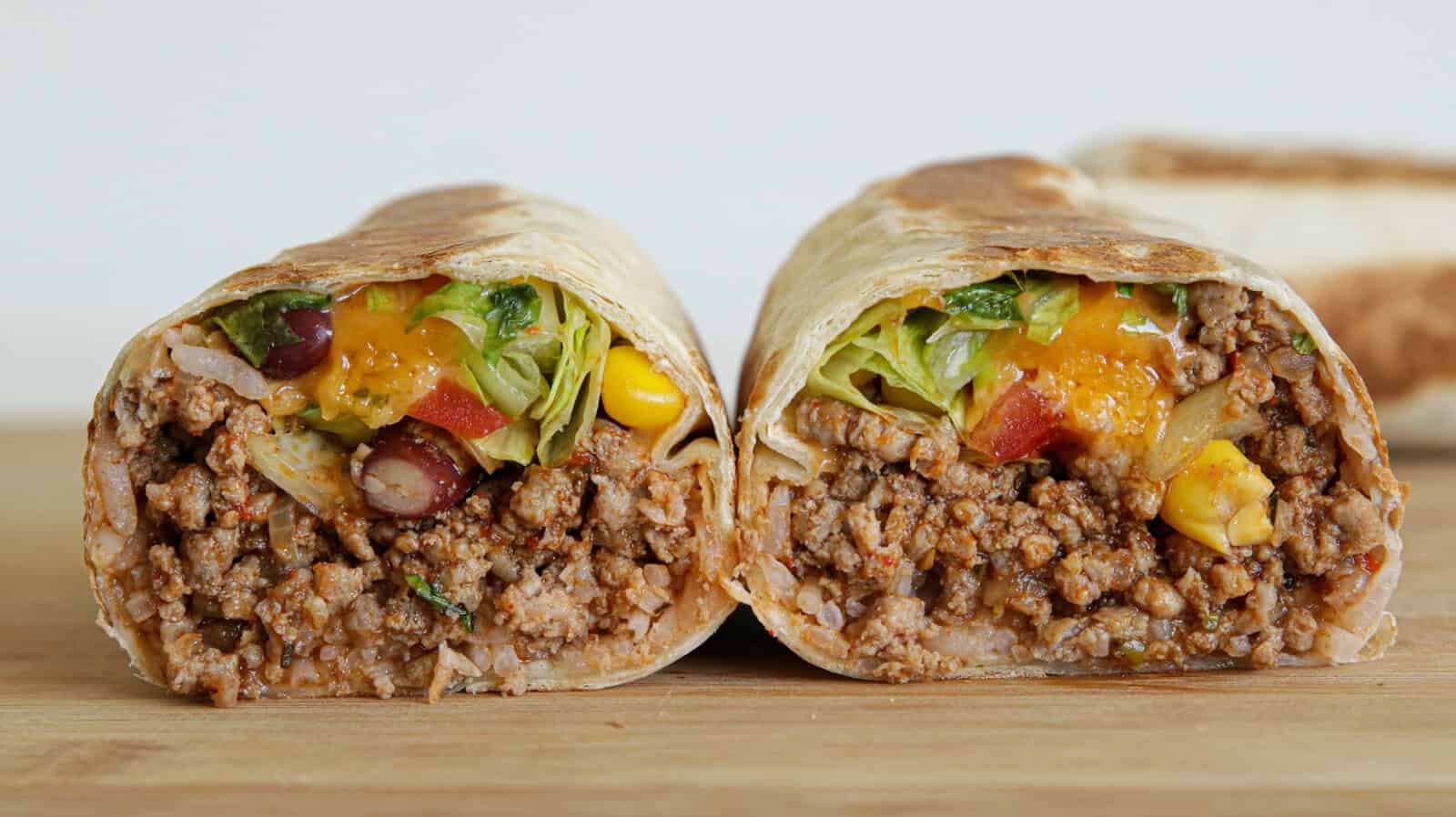 A close-up of a neatly cut burrito, showing its interior filled with ground meat, lettuce, beans, corn, and diced tomatoes. The burrito halves are placed on a wooden surface, displaying their vibrant and varied ingredients.