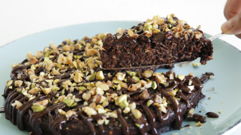 A decadent chocolate cake topped with chopped nuts and drizzled with chocolate glaze is on a light blue plate. A utensil is lifting a slice of the cake, revealing its moist interior.