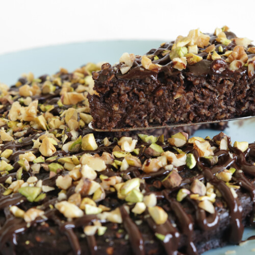 A decadent chocolate cake topped with chopped nuts and drizzled with chocolate glaze is on a light blue plate. A utensil is lifting a slice of the cake, revealing its moist interior.