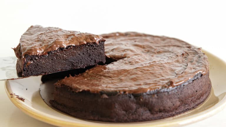 A rich, dense chocolate cake with one triangular slice being lifted out on a metal spatula. The cake has a glossy, chocolatey surface and appears moist and decadent, sitting on a light-colored plate.