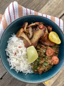 A blue bowl with white rice, roasted mushrooms, eggplant curry topped with chopped herbs and cherry tomatoes, a yellow tomato slice, a lime wedge, and red chili slices, placed on a striped cloth over a wooden surface.