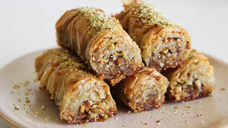 A plate of rolled baklava pastries, filled with chopped nuts and topped with a drizzle of honey and a sprinkling of ground pistachios. The layers of flaky, golden-brown phyllo dough are visible, showcasing a crunchy and syrupy texture.