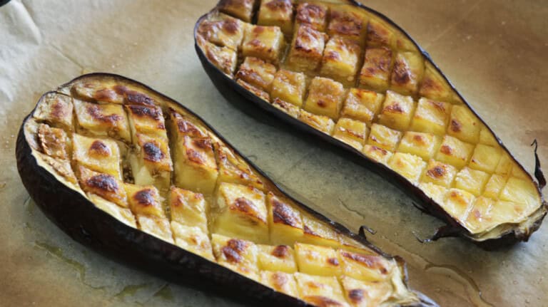 Two halves of roasted eggplant with a golden-brown, cross-hatch pattern on their flesh, placed on a baking sheet lined with parchment paper. The eggplant is cooked until soft and caramelized, with a slightly charred outer skin.