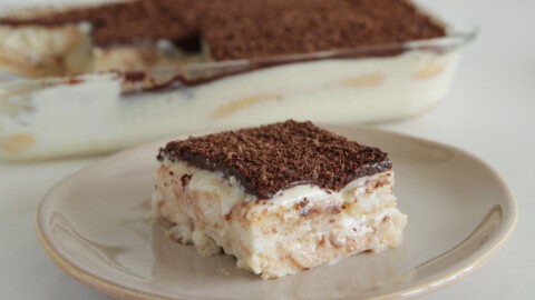 A slice of layered dessert featuring a creamy filling and a cocoa powder topping is served on a beige plate. The dessert dish with the remaining portion is visible in the background. The slice showcases distinct layers of filling and chocolate topping.