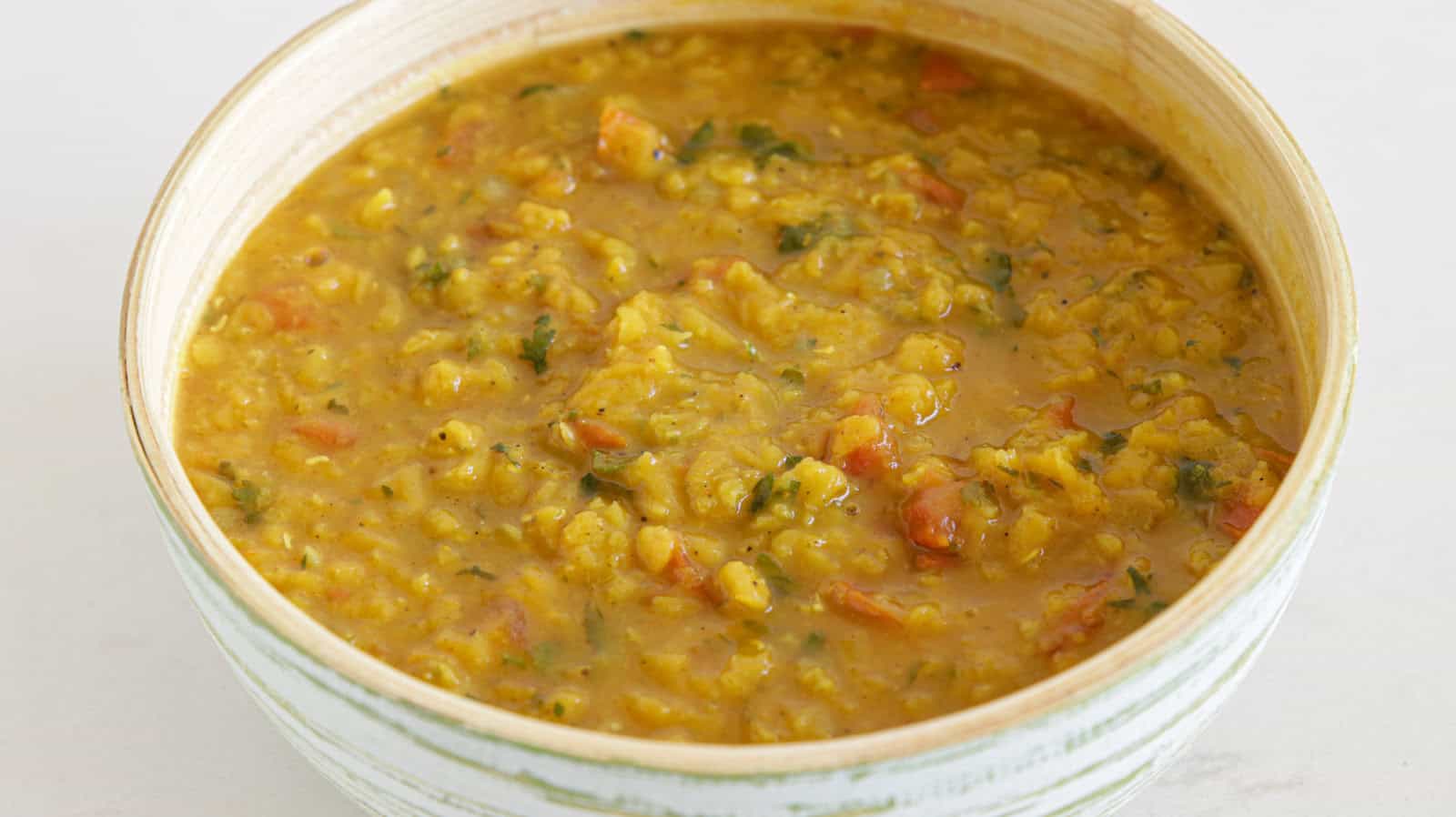 A ceramic bowl filled with a dish of yellow lentil curry. The curry appears thick and hearty, with visible pieces of lentils, tomatoes, and herbs, creating a textured and colorful appearance.