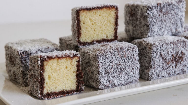 A plate of lamingtons, which are traditional Australian desserts. They are sponge cake squares coated in a layer of chocolate icing and rolled in desiccated coconut. Some of the lamingtons are cut open to reveal their soft, yellow sponge inside.