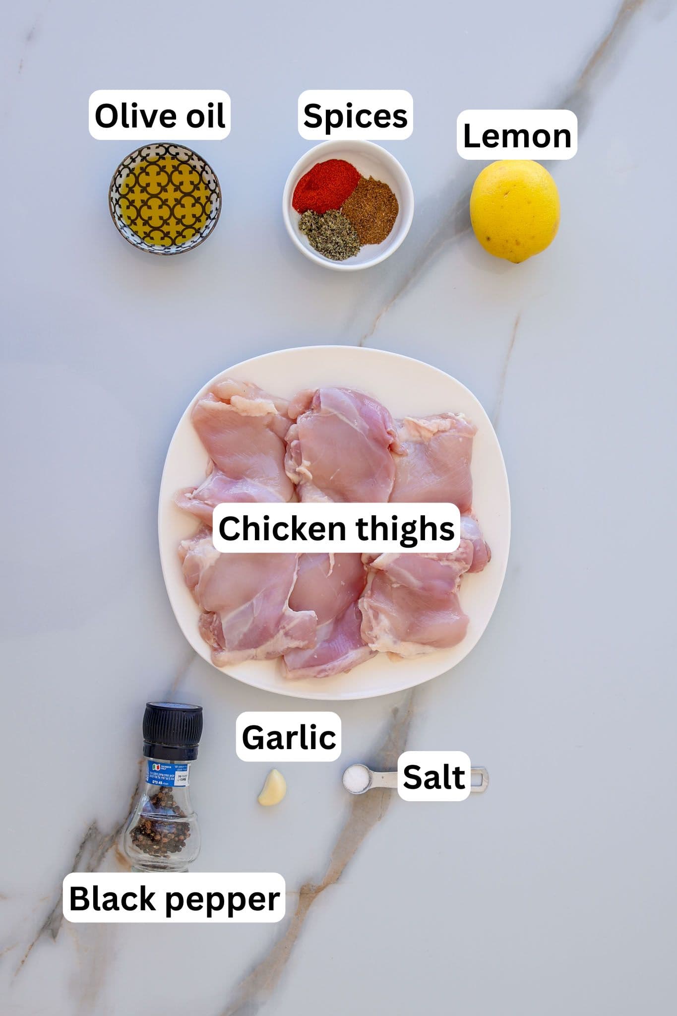 A plate of raw chicken thighs on a white surface, surrounded by labeled ingredients: a bowl of olive oil, mixed spices, a lemon, salt, black pepper, and two garlic cloves.