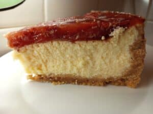 A close-up of a slice of cheesecake with a thick, creamy layer, a golden-brown crust, and a glossy red fruit topping, served on a white plate.