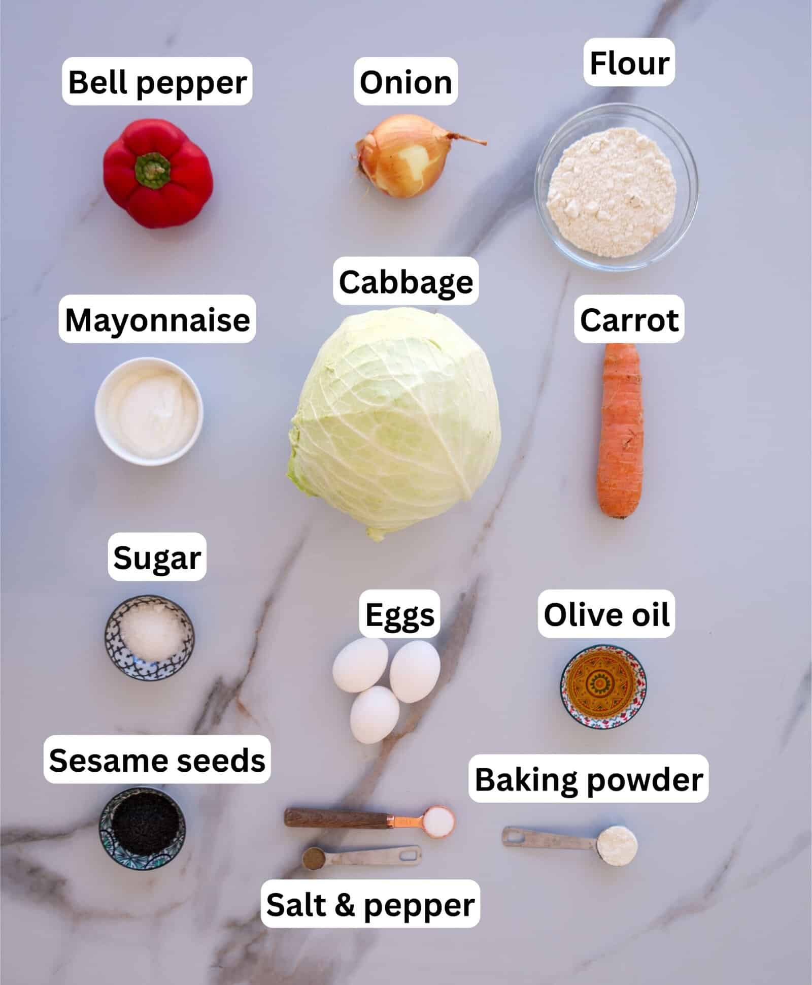 A flat lay of ingredients on a marble surface, each labeled: bell pepper, onion, flour, mayonnaise, cabbage, carrot, sugar, eggs, olive oil, sesame seeds, baking powder, salt, and pepper.