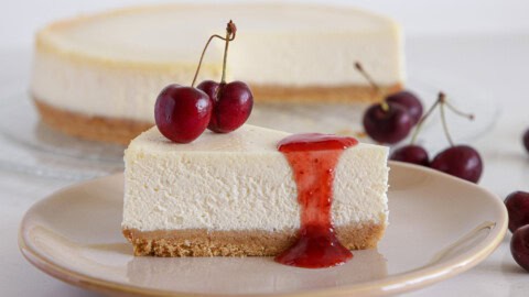 A slice of cheesecake topped with red cherry sauce and two fresh cherries on a beige plate. The creamy cheesecake has a golden brown crust. In the background, there's a whole cheesecake with a slice missing, and more cherries are scattered beside the plate.