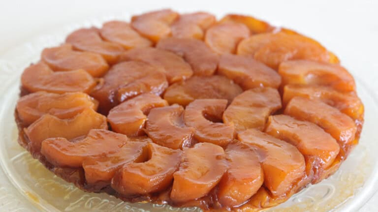 A round caramelized apple tart is displayed on a clear glass plate. The tart features neatly arranged, overlapping apple slices, golden and glossy from the caramel glaze on top. The crust appears slightly visible at the edges, hinting at a flaky pastry base.