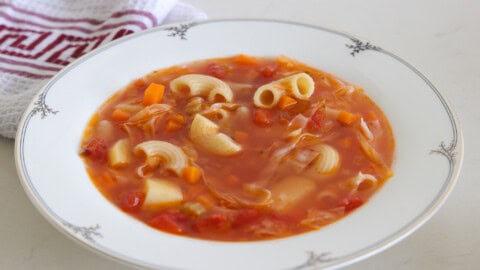 A white bowl filled with minestrone soup, featuring ingredients such as elbow pasta, diced carrots, tomatoes, and a broth, sits on a white surface. A white and red patterned cloth is partially visible in the background.