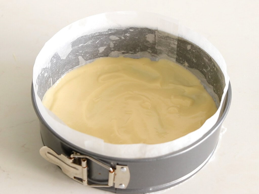 A round springform pan lined with parchment paper, filled with smooth, pale cake batter, sits on a light-colored countertop.