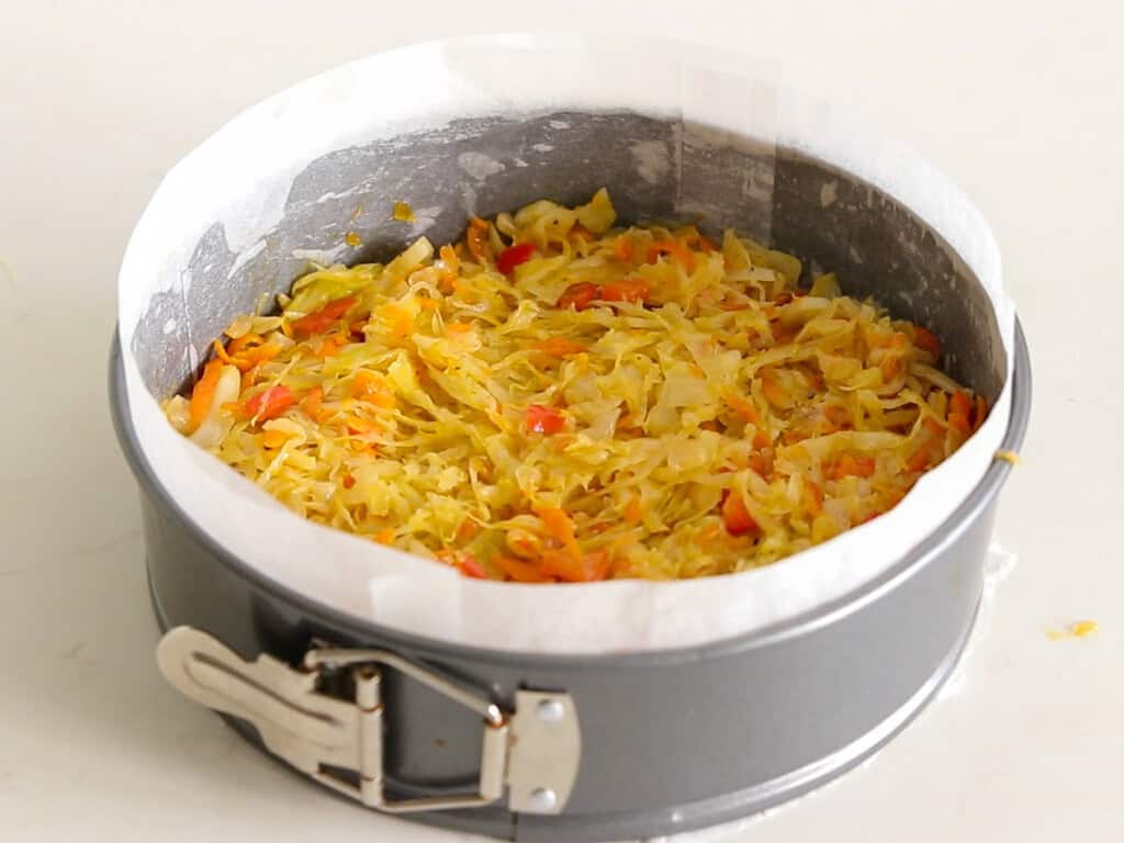 A springform pan lined with parchment paper, filled with a mixture of shredded cabbage, carrots, and red peppers, sits on a light-colored countertop.
