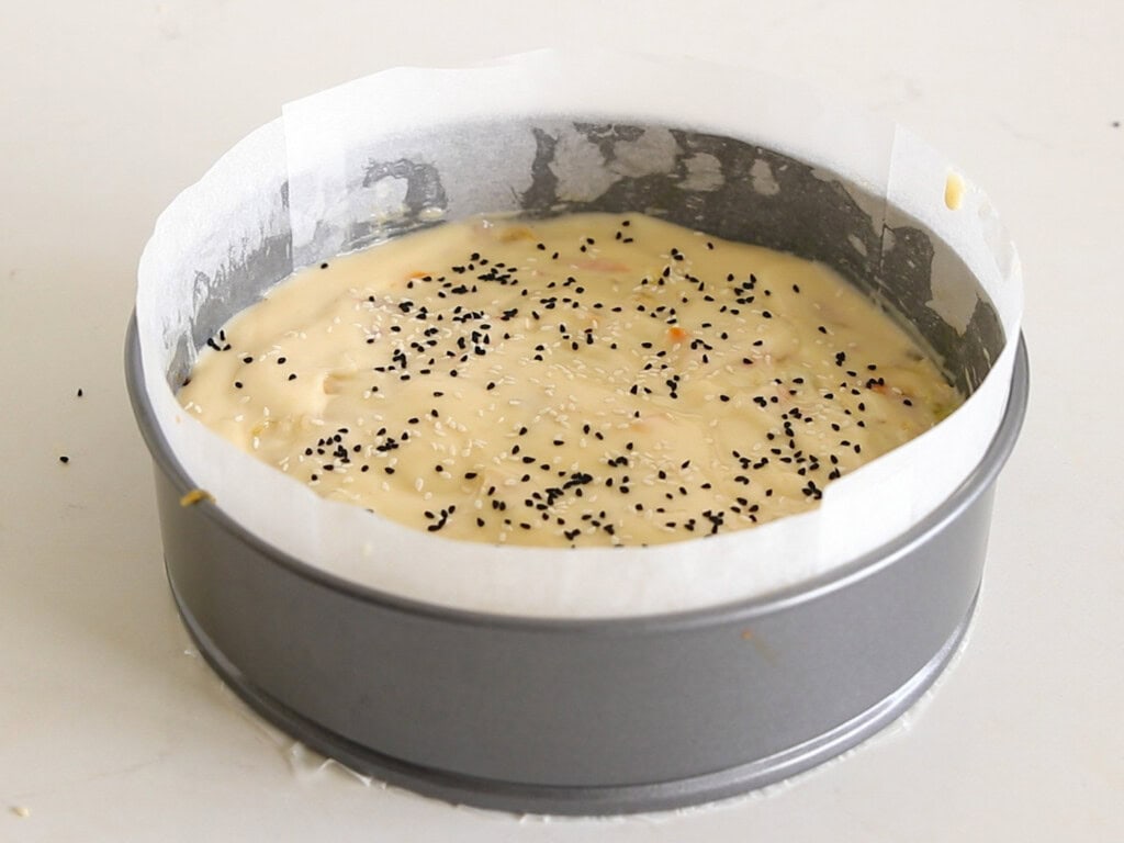 A round cake pan lined with parchment paper contains unbaked cake batter sprinkled with black seeds, sitting on a white surface.