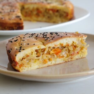 A slice of savory pie with a golden, fluffy crust, filled with shredded cabbage and carrots, topped with sesame and black seeds, sits on a plate. The rest of the pie is visible in the background.
