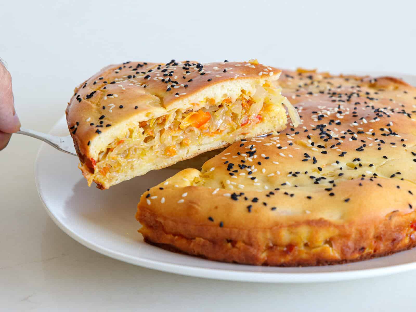 A savory Russian cabbage pie topped with sesame and nigella seeds is being sliced and served from a white plate, revealing a filling of shredded vegetables such as cabbage and carrots.