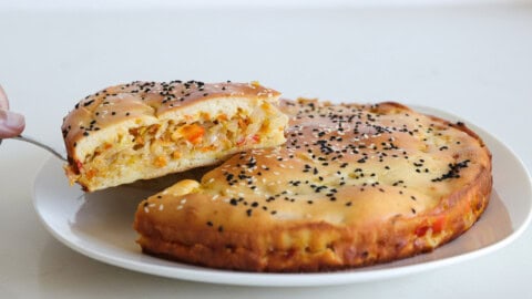 A round savory pie topped with black and white sesame seeds sits on a white plate, with a slice being lifted to show a filling of chopped vegetables.
