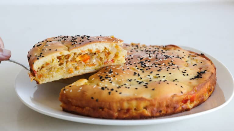 A round savory pie topped with black and white sesame seeds sits on a white plate, with a slice being lifted to show a filling of chopped vegetables.