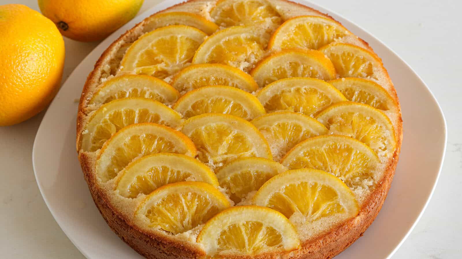 A round lemon cake on a white plate is topped with neatly arranged semi-circular slices of lemon. Two whole lemons are placed next to the plate on a light-colored surface.