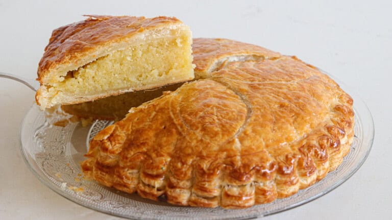 A round Galette des Rois, a traditional French cake with a flaky, golden-brown crust, is displayed on a glass plate. A slice has been cut and is slightly lifted, revealing a dense, almond frangipane filling inside.