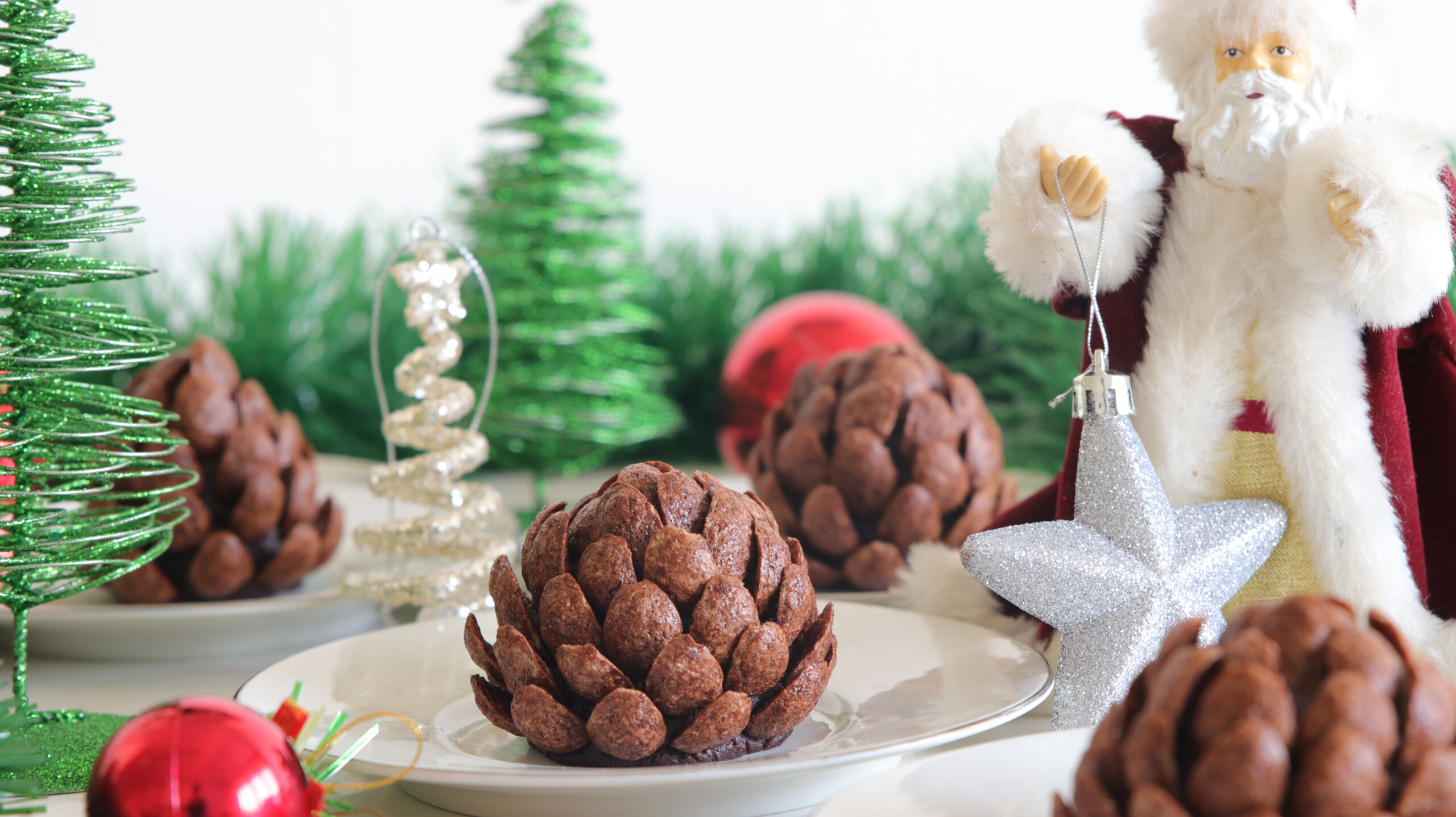 A festive scene featuring chocolate pine cone desserts on a white plate, surrounded by Christmas ornaments and decorations. There is a Santa figurine holding a silver star ornament, with green tinsel and Christmas trees in the background.