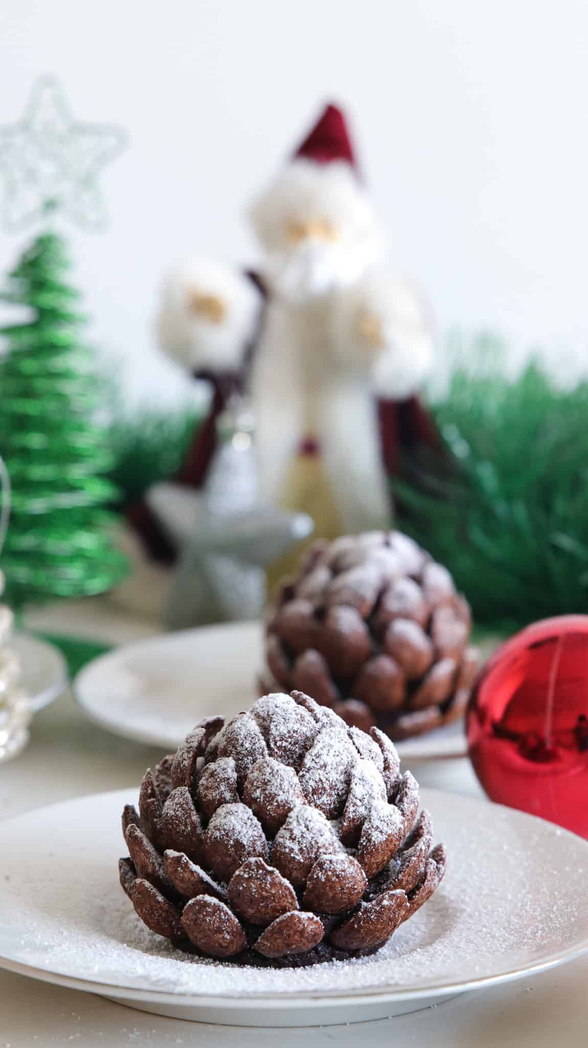 A festive scene featuring chocolate pine cone desserts on a white plate, surrounded by Christmas ornaments and decorations. There is a Santa figurine holding a silver star ornament, with green tinsel and Christmas trees in the background.