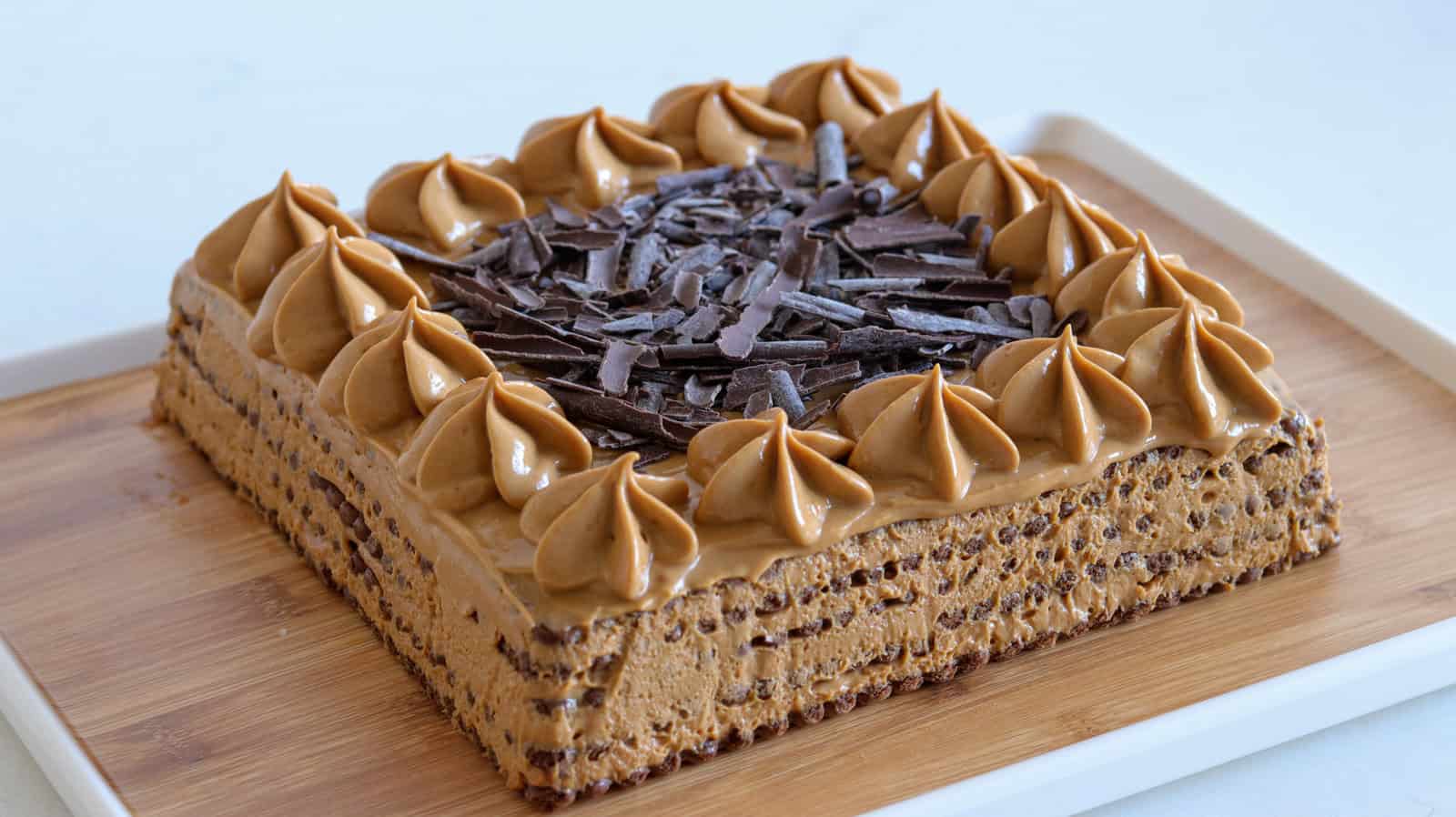 A square chocotorta cake topped with rosettes of coffee frosting and a generous amount of chocolate shavings in the center. The cake sits on a wooden board, with a white background.