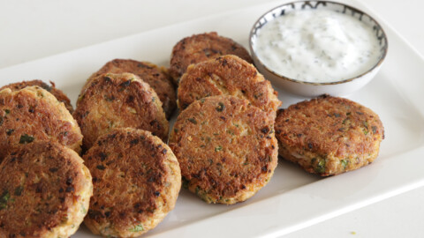 A rectangular white plate holds several round, golden-brown tuna patties with a crispy texture. A small bowl filled with white tzatziki sauce garnished with herbs is placed on the plate next to the tuna patties.