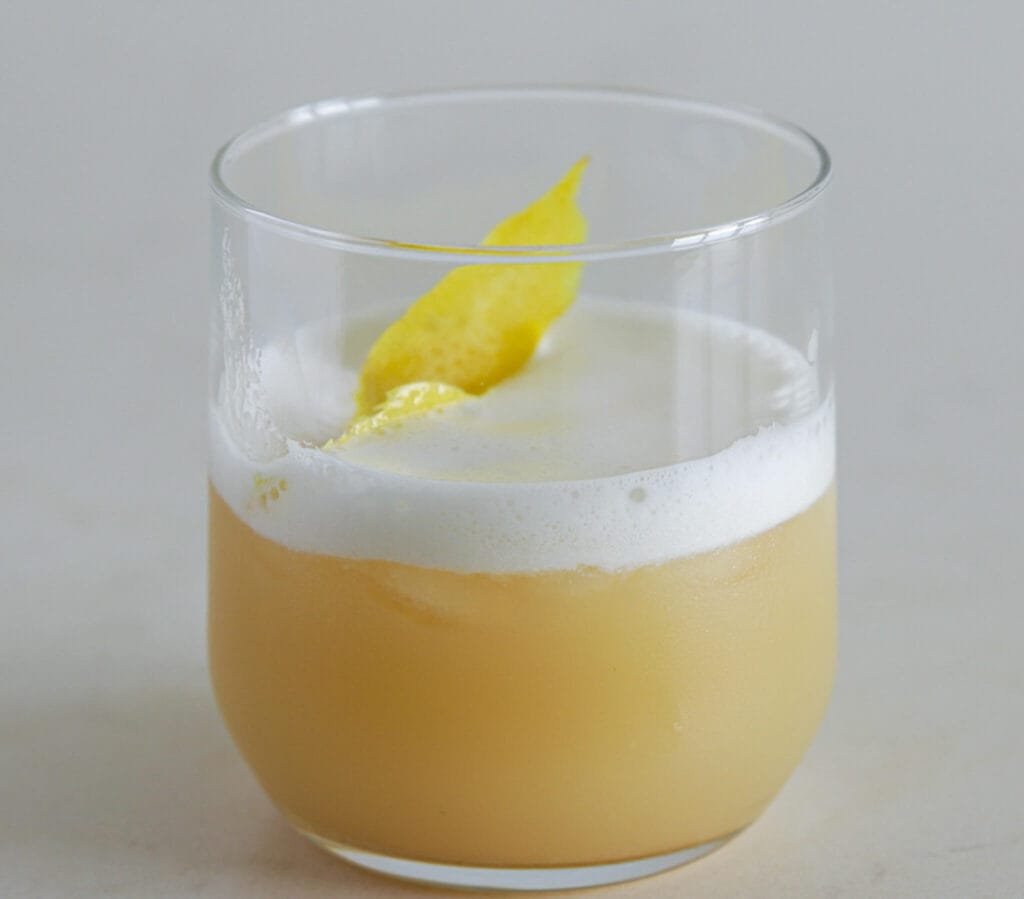 A transparent glass filled with a frothy, light beige drink garnished with a lemon peel. The beverage has a creamy top layer and is set against a simple white background.