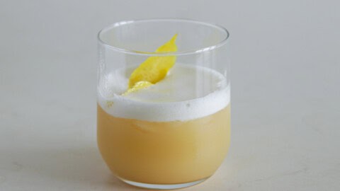 A cocktail in a clear glass, featuring a frothy white layer on top and a pale peach-colored liquid below. The drink is garnished with a twist of lemon peel, which rests on the foam. The background is plain and light-colored.