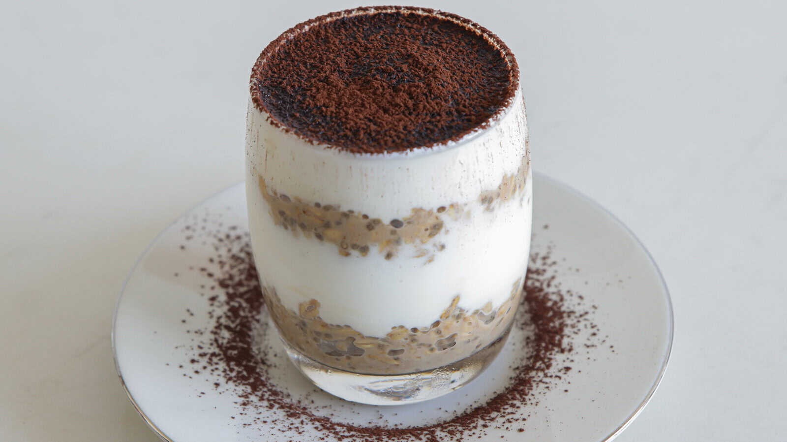 overnoght layered in a transparent glass is placed on a white plate. The dessert has visible layers of creamy white substance and a brown ingredient, possibly cocoa or coffee powder, sprinkled on top. There is also some powdered topping on the plate.