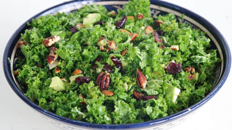 A close-up of a bowl filled with fresh kale salad. The salad contains chopped kale leaves, avocado pieces, dried cranberries, pecan nuts, and sliced almonds, all tossed together. The bowl has an intricately patterned blue and white border.