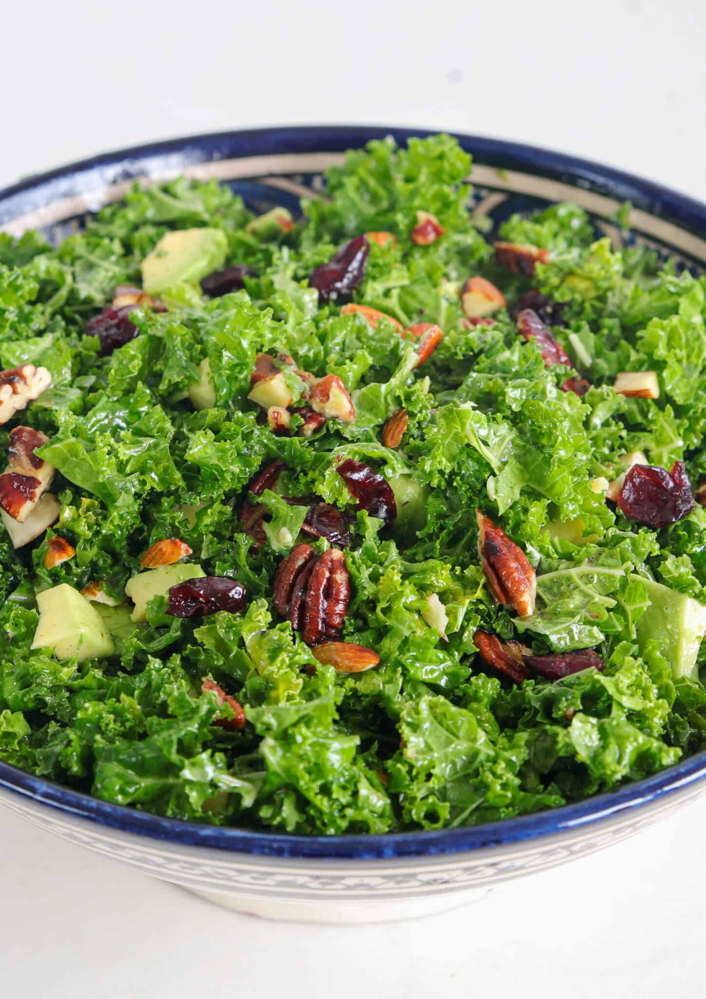 A close-up view of a fresh kale salad in a blue-patterned bowl. The salad contains chopped avocado, dried cranberries, pecans, and a mix of herbs, all tossed together and glistening as if lightly dressed.