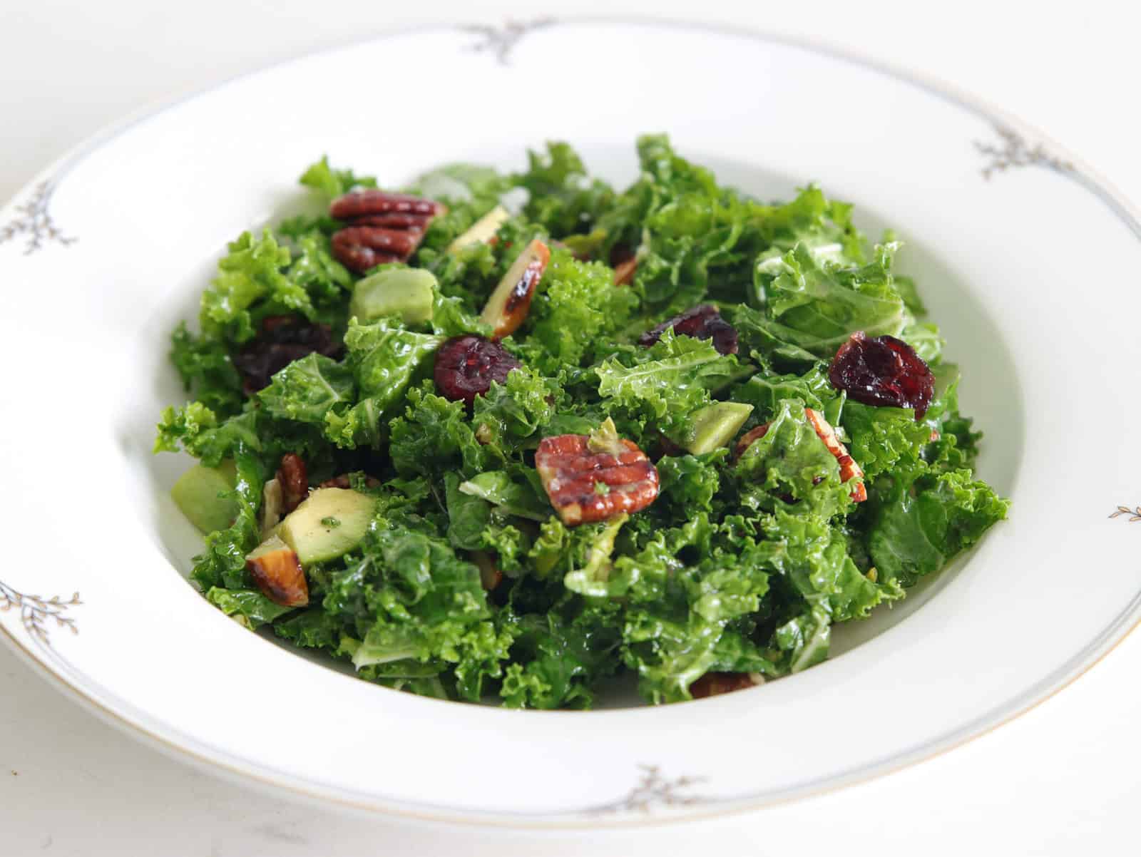 A white decorative bowl filled with a fresh kale salad, featuring chopped avocados, red dried cranberries, and pecans. The salad ingredients are vibrantly green and well-mixed, creating a colorful and healthy presentation.