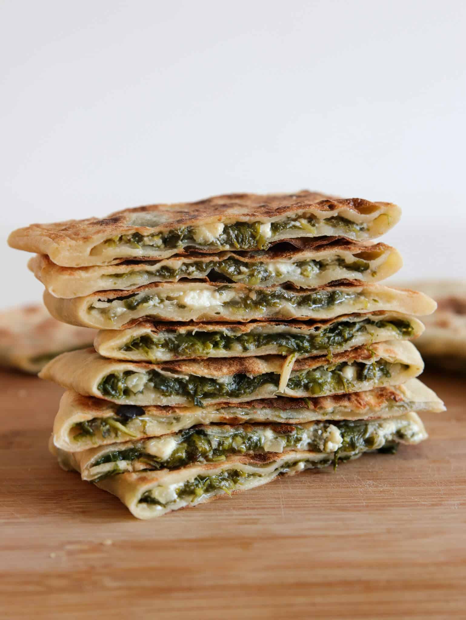 A stack of Turkish gozleme with a green and white filling, likely spinach and cheese, cut in half to show the layers, placed on a wooden surface with a light background.