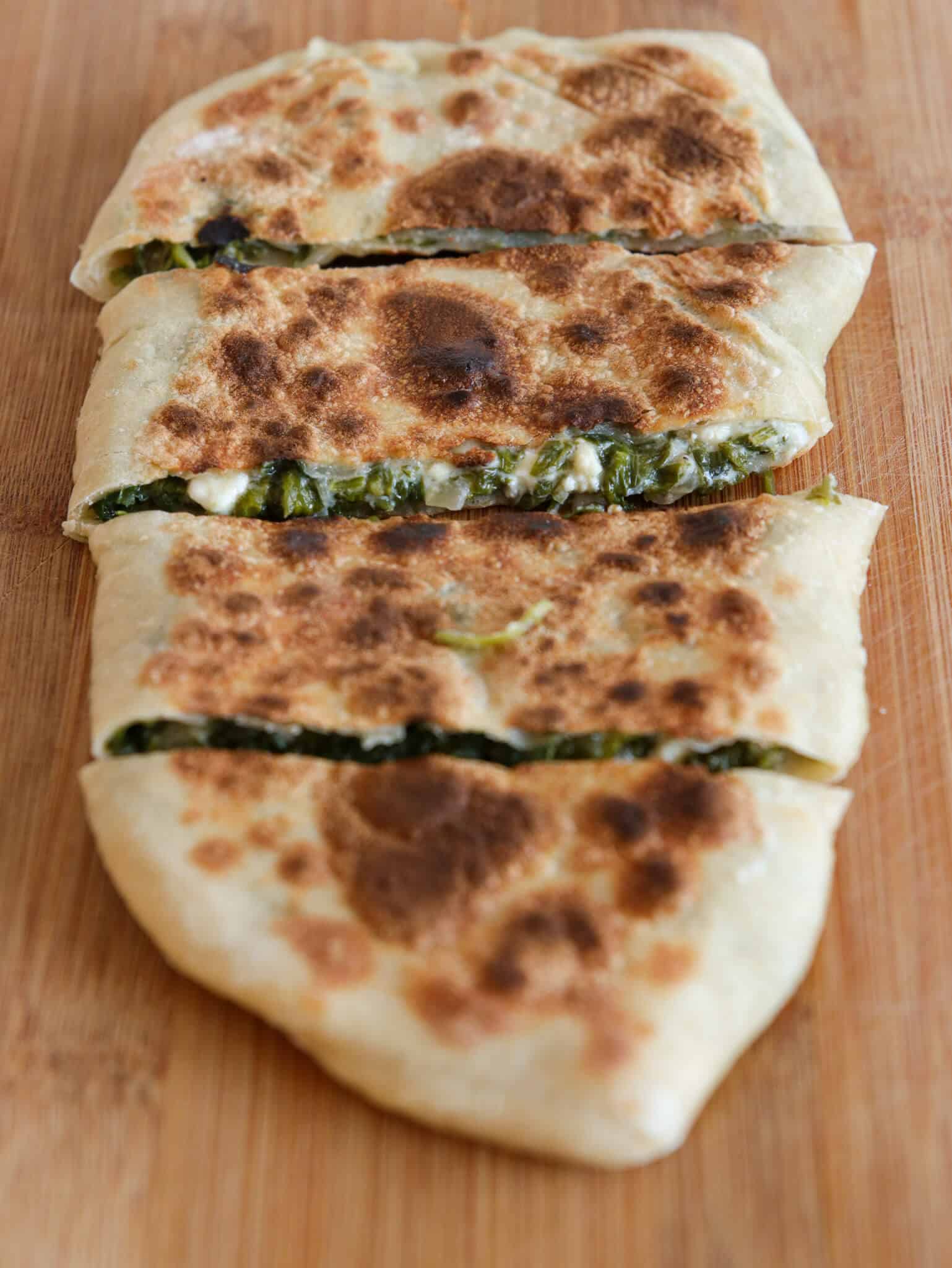 A sliced, golden-brown flatbread filled with cooked greens and cheese, displayed on a wooden cutting board.