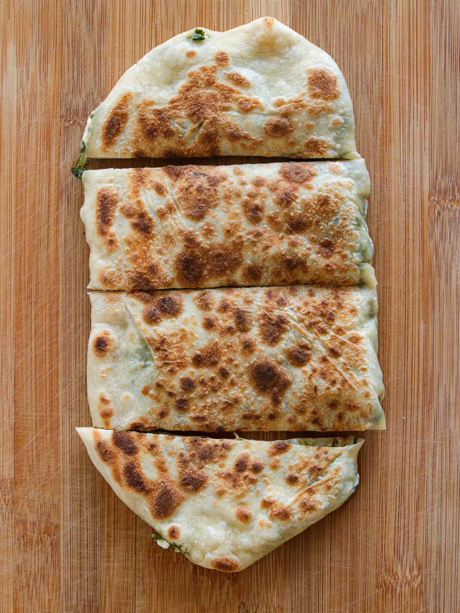 A golden-brown stuffed flatbread, sliced into four pieces, sits on a wooden cutting board. The bread has a crisp, toasted surface with darker spots and slightly uneven edges.
