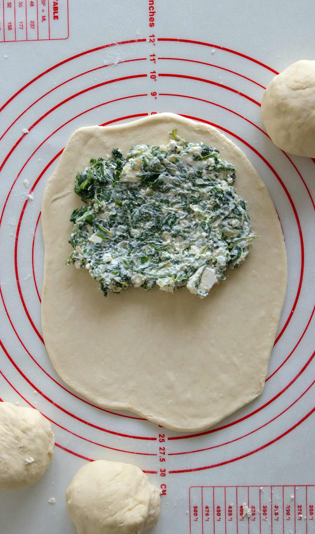 A rectangle of rolled dough on a red and white pastry mat, topped with a spinach and cheese mixture. Three dough balls rest nearby, ready to be filled.